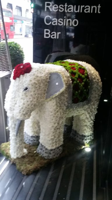 Textured elephant sculpture adorned with flowers, displayed in a shop window.
